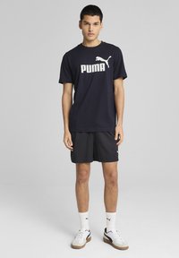 Marineblauwe t-shirt met wit "PUMA" logo, gecombineerd met zwarte shorts. Het model draagt witte en grijze sneakers. Eenvoudig, sportief ontwerp.