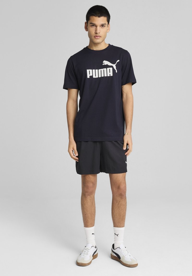 Námořnické tričko s bílým logem "PUMA", v kombinaci s černými šortkami. Model má na sobě bílé a šedé tenisky. Jednoduchý, sportovní design.
