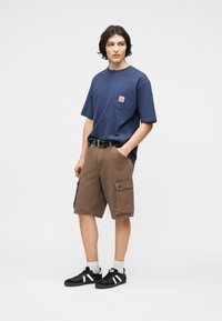 Άνδρας που φοράει μπλε σκούρο μπλουζάκι, καφέ cargo σορτς με τσέπες, μαύρη ζώνη, λευκές κάλτσες και μαύρα αθλητικά παπούτσια, στέκεται με το ένα χέρι στην τσέπη.