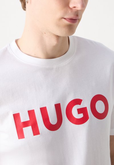 Бяла памучна тениска сBold червен текст, който изписва "HUGO." Класически кръгъл вратник с гладка текстура и къси ръкави.