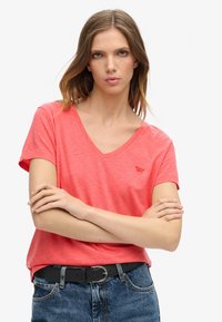 Korallfärgad V-hals t-shirt i mjukt, lätt material, med korta ärmar och en subtil logotyp på bröstet. Kombinerad med högmidjade jeans.