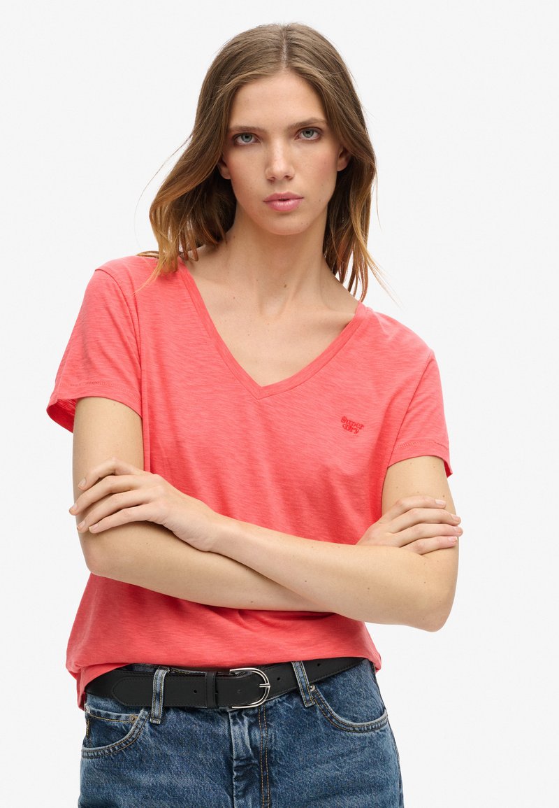 Mujer joven con cabello castaño claro, vestida con una camiseta coral de cuello en V y jeans azules, con los brazos cruzados y expresión neutral.