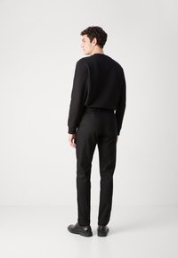 Chemise noire à manches longues, en tissu texturé, assortie à un pantalon noir ajusté. Des chaussures noires complètent la tenue, vue de dos.