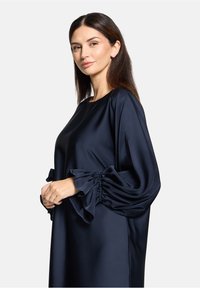 Robe en satin bleu marine avec une coupe ample, dotée de manches bouffantes avec des détails froncés aux poignets. Texture lisse, design minimaliste.