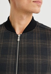 Muž v tmavě kostkované bundě s černým žebrovaným límcem a stříbrným zipem, s hlavou mírně otočenou na stranu.