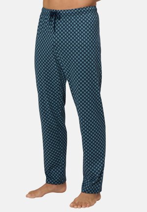 Mann trägt blaue gemusterte Pyjamahose mit Kordelzug am Bund, steht barfuß vor schlichter Hintergrund.