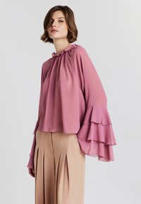 Donna che indossa una blusa rosa fluida con maniche a campana a strati e pantaloni beige a vita alta con pieghe, su uno sfondo neutro.
