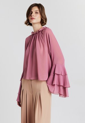 Donna che indossa una blusa rosa fluida con maniche a campana a strati e pantaloni beige a vita alta con pieghe, su uno sfondo neutro.