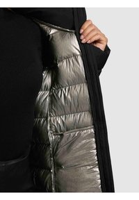 Kvinde iført sort sweater viser sølvfarvet polstret foring inde i en sort quiltet jakke ved at holde den åben med sin højre hånd.