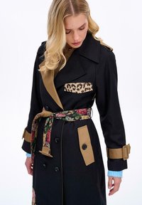 Czarny płaszcz typu trench z kieszenią na piersi w panterkę, beżowymi akcentami i paskiem w kwiatowy wzór. Charakteryzuje się dużymi guzikami i stylowym kołnierzem.