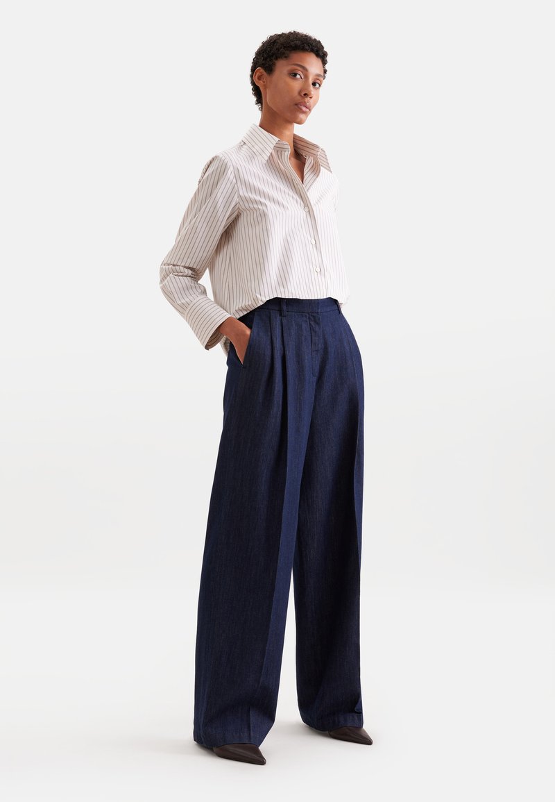 Femme portant une chemise légère rayée boutonnée et un pantalon bleu foncé taille haute à jambes larges, les mains dans les poches.