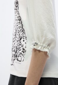 Τοπ ανοιχτού χρώματος με γραφικό σχέδιο λεοπάρδαλης. Διαθέτει φουσκωτά μανίκια με ελαστ ικά τελειώματα και διακριτική λάμψη στην υφή του υφάσματος.