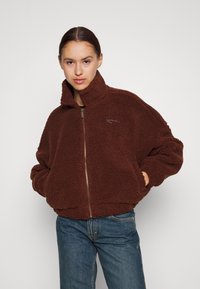 Brun fleecejakke med høy krage, glidelås foran og elastiske mansjetter. Har et teksturert, fluffy materiale og diskret logo-broderi.