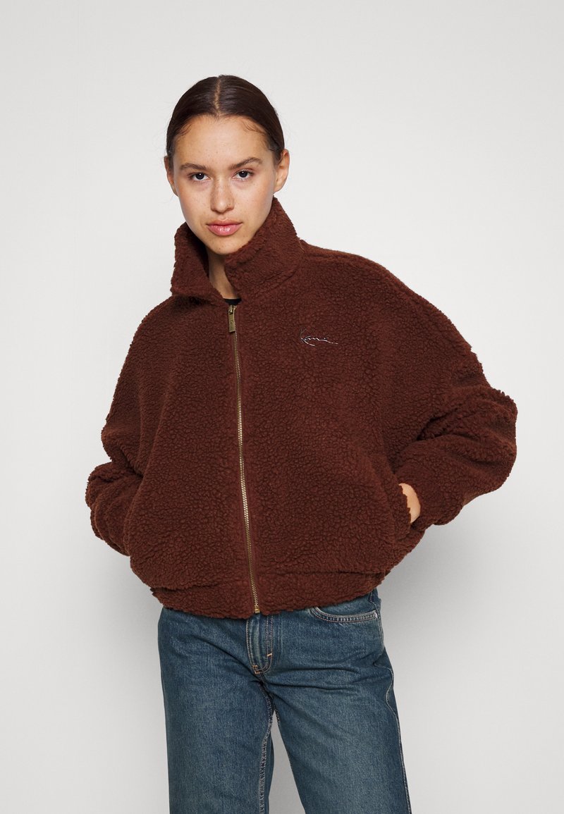 Brun fleecejakke med høy krage, glidelås foran og elastiske mansjetter. Har et teksturert, fluffy materiale og diskret logo-broderi.