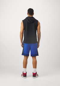 Fekete ujjatlan kapucnis felső kék csíkos sorttal, fekete oldalsó panelekkel. Piros-fehér sportcipők, látható fehér zoknikkal.