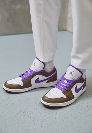 Baskets blanches et marron avec des lacets violets et un logo, portées avec un pantalon blanc et des chaussettes blanches sur un sol bleu clair tacheté.