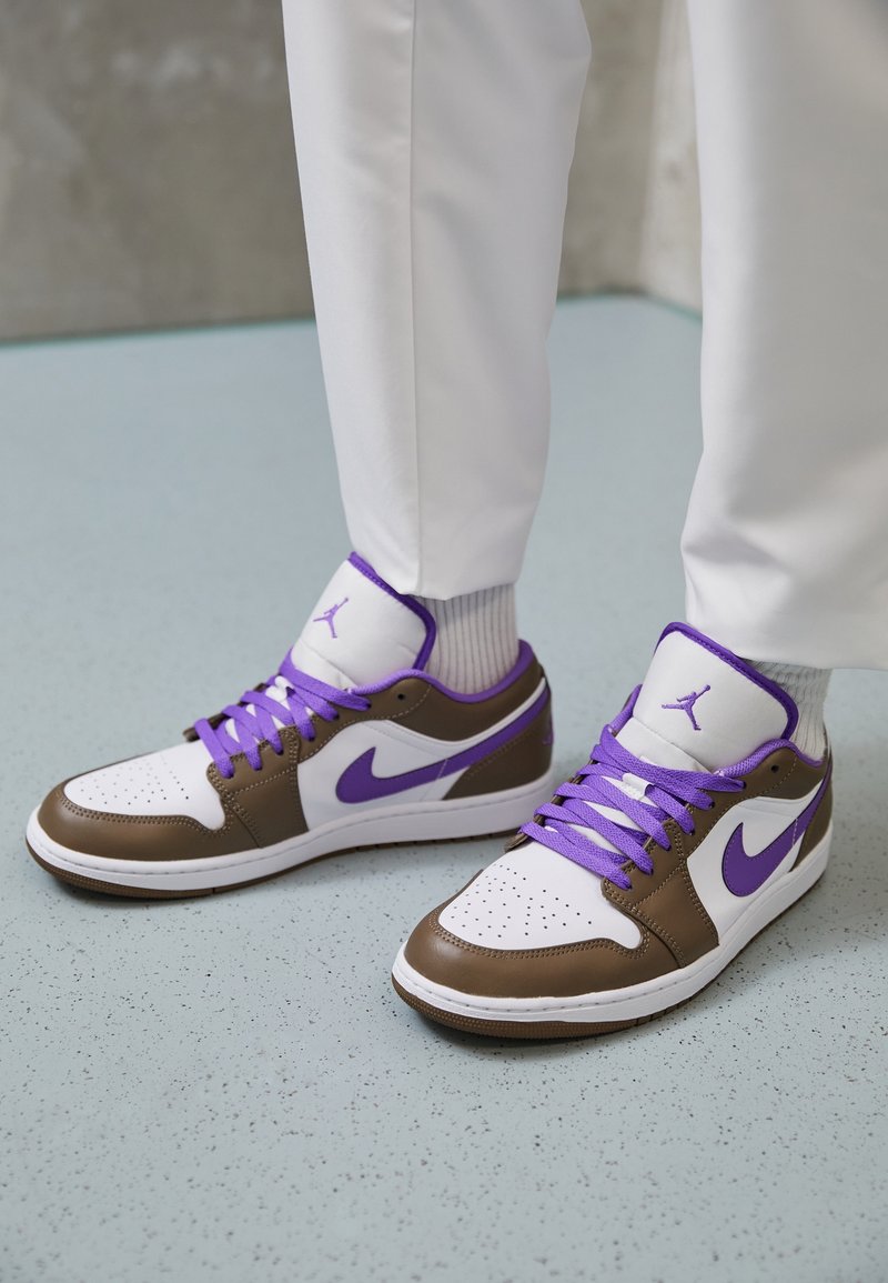 Baskets blanches et marron avec des lacets violets et un logo, portées avec un pantalon blanc et des chaussettes blanches sur un sol bleu clair tacheté.