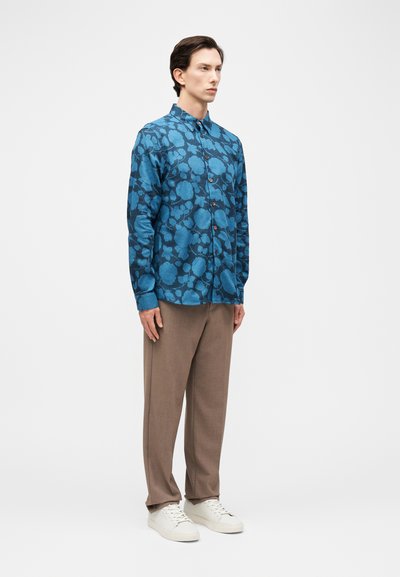 Άνδρας που στέκεται, φοράει μπλε πουκάμισο με λουλουδένιο σχέδιο και μακριά μανίκια, καφέ παντελόνια και λευκά αθλητικά παπούτσια σε απλό φωτεινό φόντο.