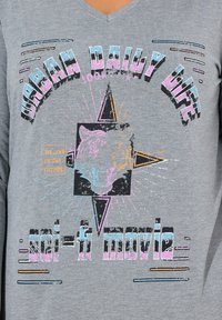 Grijze V-hals t-shirt met een grafisch ontwerp van een tijgerkop, vetgedrukte tekst en kleurrijke accenten in blauw, roze en oranje. Verweerde uitstraling.