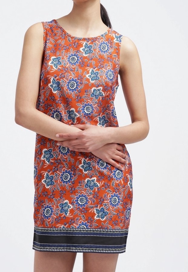 Femme portant une robe sans manches orange avec un motif floral bleu et blanc, les bras croisés, debout devant un fond blanc uni.