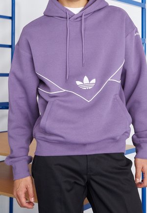 Mężczyzna ubrany w fioletową bluzę z kapturem Adidas z białym wykończeniem i logo, w czarnych spodniach, stoi w pomieszczeniu blisko niebieskich poręczy.