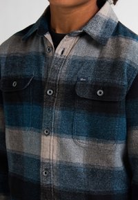 Camisa de franela con un audaz patrón de cuadros en azul, negro y gris, cierre de botones, dos bolsillos en el pecho y un tejido suave y texturizado.