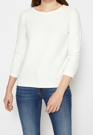 Weißer strukturierter Pullover mit rundem Ausschnitt und langen Ärmeln, mit gerafften Details an den Manschetten, kombiniert mit blauen Jeans.