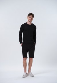 Schwarzes Langarmshirt mit dezentem Muster, kombiniert mit schwarzen Shorts und hellgrauen Schuhen. Minimalistisches Design und neutrale Farbgebung.