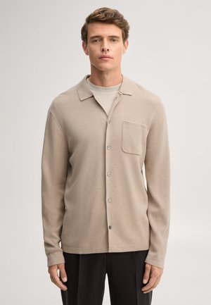 Junger Mann, der einen beigen, geknöpften Cardigan über einem hellen Hemd und schwarzen Hosen trägt, vor einem schlichten hellen Hintergrund stehend.