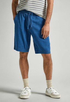 Man draagt blauwe shorts met koord, beige sokken, witte sneakers en een gestreept overhemd, met één hand in de zak en één ontspannen langs het lichaam.