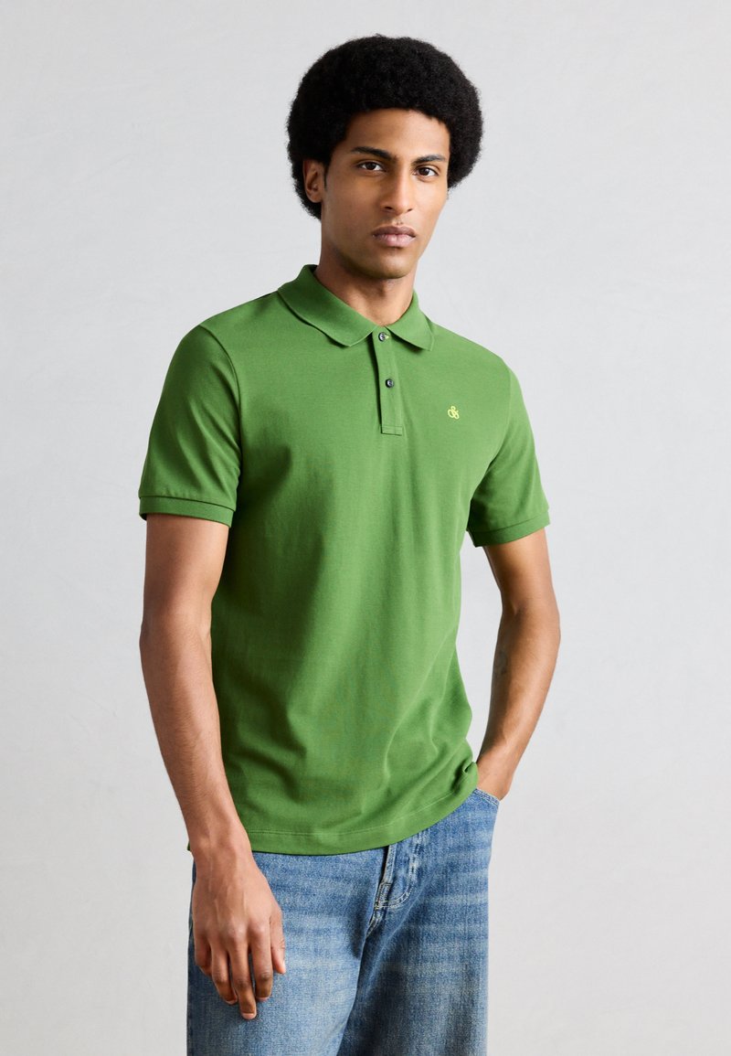 Mann mit Afro-Frisur, der ein grünes Poloshirt und blaue Jeans trägt und vor einem einfachen hellgrauen Hintergrund steht.