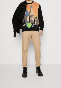 Man i svart och orange tröja med flaska- och tärningsdesign, beige byxor, svarta stövlar, som håller en svart jacka över axeln mot vit bakgrund.