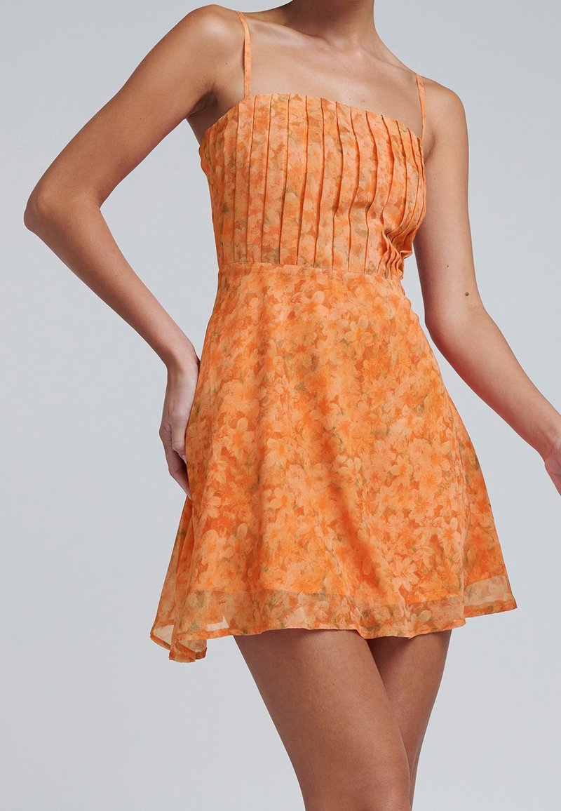 Femme portant une robe trapèze fleurie orange avec fines bretelles et un corsage plissé sur un fond gris uni.