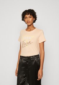 Lichtroze katoenen t-shirt met een ronde halslijn, voorzien van een gele driehoek en glanzend logo detail in het midden. Gecombineerd met glanzende zwarte broeken.