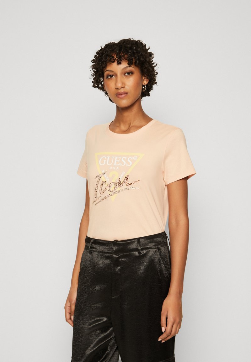 Lichtroze katoenen t-shirt met een ronde halslijn, voorzien van een gele driehoek en glanzend logo detail in het midden. Gecombineerd met glanzende zwarte broeken.