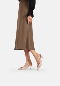 Slingback di colore rosa chiaro con punta appuntita, tacco basso e dettaglio decorativo a fiocco. Texture in pelle liscia, completata da una cintura regolabile.