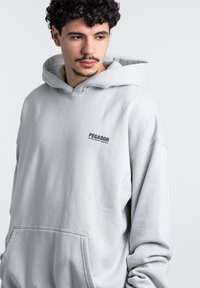 Felpa grigia chiara con cappuccio e tasca frontale, con il logo "PEGADOR" in nero. Tessuto liscio, vestibilità comoda e maniche larghe.