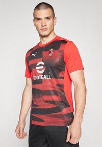 Camiseta de fútbol de manga corta en rojo y negro con un patrón de rayas horizontales, logo de Puma y emblema del equipo en el pecho. Tejido ligero.