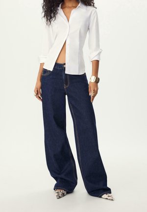 Kvinde iført en delvist knappet hvid skjorte, vidde mørkeblå jeans, leopardmønstrede spidse sko og guldsmykker på en hvid baggrund.