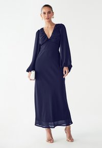 Tmavě modré šaty s dlouhým rukávem, hlubokým výstřihem do V, nařaseným materiálem na hrudi a splývavou sukní. Kombinovány se zlatými sandály na pásky a bílou kabelkou.