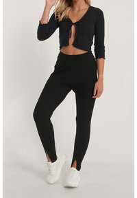 Svart ribbad crop top med knytband framtill och trekvartslånga ärmar, matchad med svarta ribbade leggings med ankelslitsar. Vita sneakers.