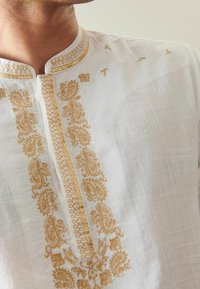 Primo piano di una persona che indossa un indumento bianco con intricati ricami dorati attorno al colletto e alla patta anteriore.