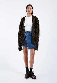 Cardigan nero e oro con texture, di taglia over, con bottoni, indossato sopra un top bianco aderente e una gonna di jeans. Calze nere e scarpe scure completano l'outfit.