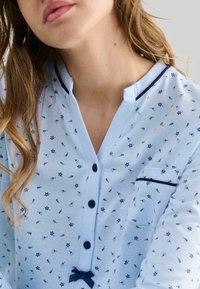 Primer plano de una persona con una camisa de botones floral en azul claro, con botones azul oscuro y un pequeño bolsillo en el pecho.