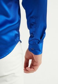 Chemise en satin bleu à manches longues, avec une coupe ajustée, des poignets boutonnés et une texture lisse, associée à un pantalon blanc.