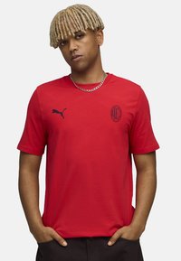 Rød kortærmet t-shirt lavet af bomuld med rund halsudskæring, prydet med et marineblåt Puma-logo og et rundt AC Milan-emblem på brystet.