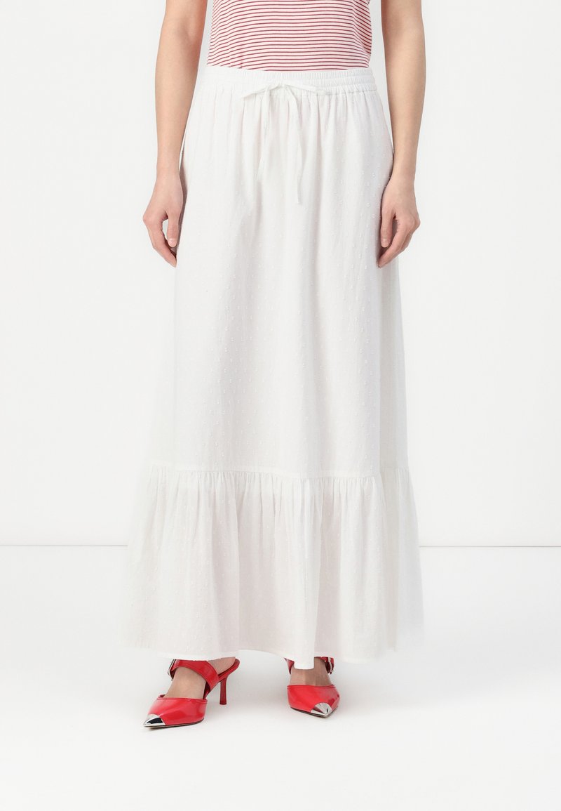 Jupe maxi blanche en tissu léger, dotée d'un ourlet à volants à étages et d'une texture verticale, associée à des chaussures à talons hauts rouges.