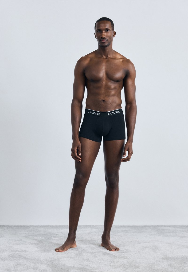 Black cotton boxer shorts with a wide elastic waistband featuring the Lacoste logo. Model is standing barefoot against a neutral background.