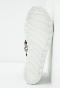 Semelle de sandale avec motif de bande de roulement texturé blanc et sangles grises visibles sur fond blanc, positionnée verticalement contre une surface blanche.