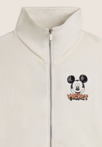 Sweat à zip blanc avec col haut, ornée d'un graphique noir et blanc de Mickey Mouse et du texte "MICKEY" en orange. Texture de tissu lisse.